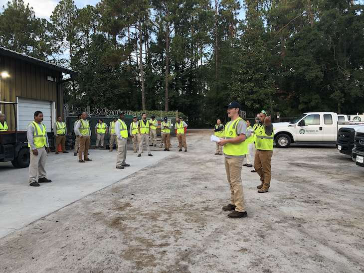 Landscape professionals listen to a speech while wearing safety vests and standing outdoors